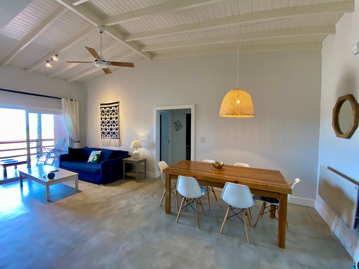 An open living and dining area features high, white-beamed ceilings. A wooden dining table with six chairs occupies the center, while a blue sofa is positioned nearby. A ceiling fan adds comfort, and large windows offer natural light and a view of the outdoors.