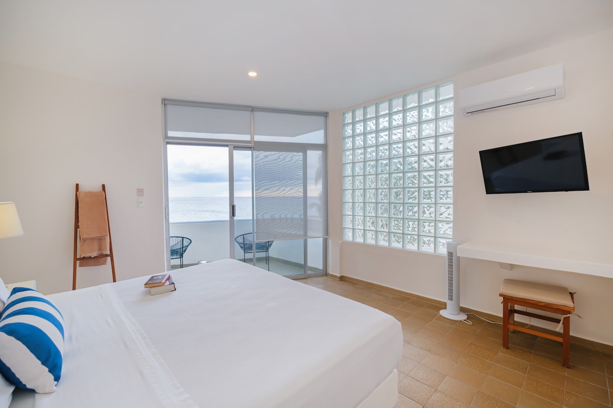 A spacious bedroom features a king-size bed with a striped blue and white pillow. Large glass doors provide a view of the ocean and lead to a private terrace. A flat-screen TV and a wooden desk are visible, while natural light enters through decorative glass blocks.