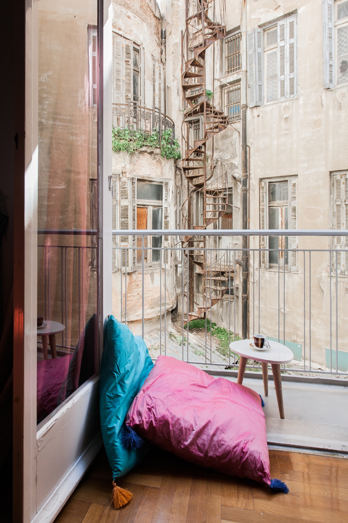 A small balcony is featured with vibrant cushions in pink and turquoise. A wooden table sits adjacent to the wall, while a spiral staircase can be seen leading to upper levels of the building, surrounded by aged walls and windows that reflect a vintage aesthetic.