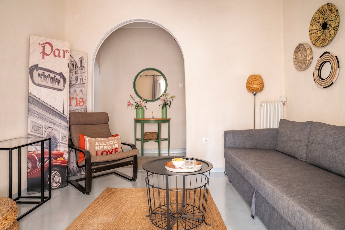 A spacious living area features a blend of comfort and style, including a modern grey sofa and a wicker chair. A round coffee table sits in the center, accented by decorative items. Natural light illuminates the room, creating a warm atmosphere.