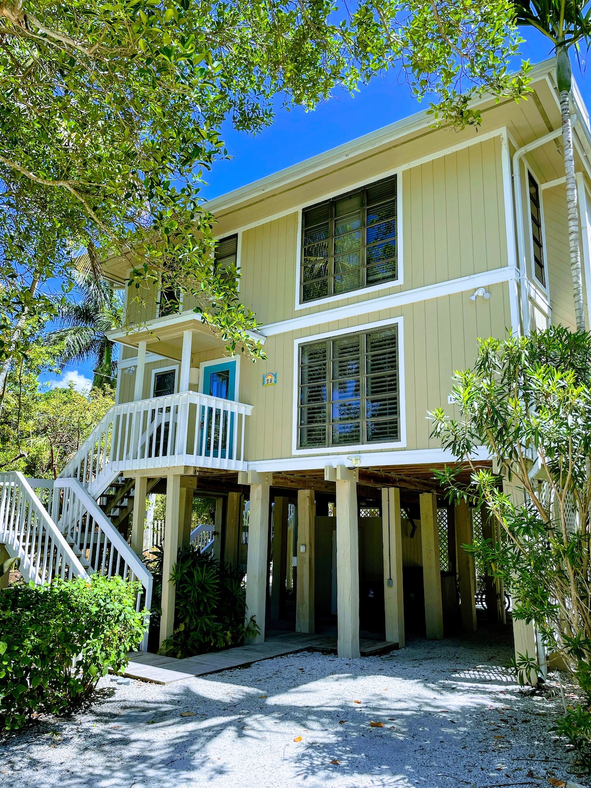 A two-story home is framed by lush greenery, showcasing a light yellow exterior. The property features large windows and a bright blue door, while a covered entryway is supported by white columns. Outdoor stairs lead to the main entrance, enhancing accessibility.