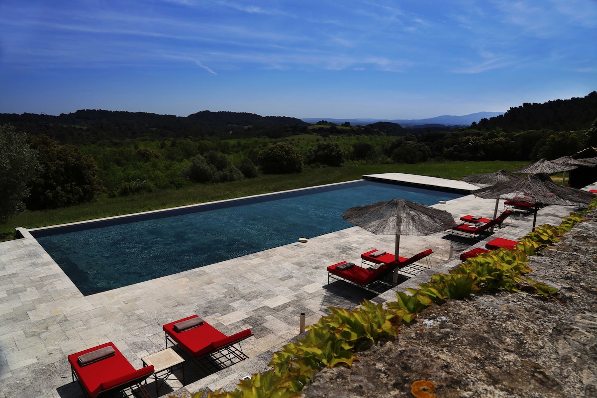 A swimming pool is situated in a serene outdoor setting, framed by lush greenery and rolling hills. Comfortable red lounge chairs are positioned beside the pool, complemented by shaded umbrellas. The clear water reflects the blue sky, enhancing the tranquil ambiance of the location.