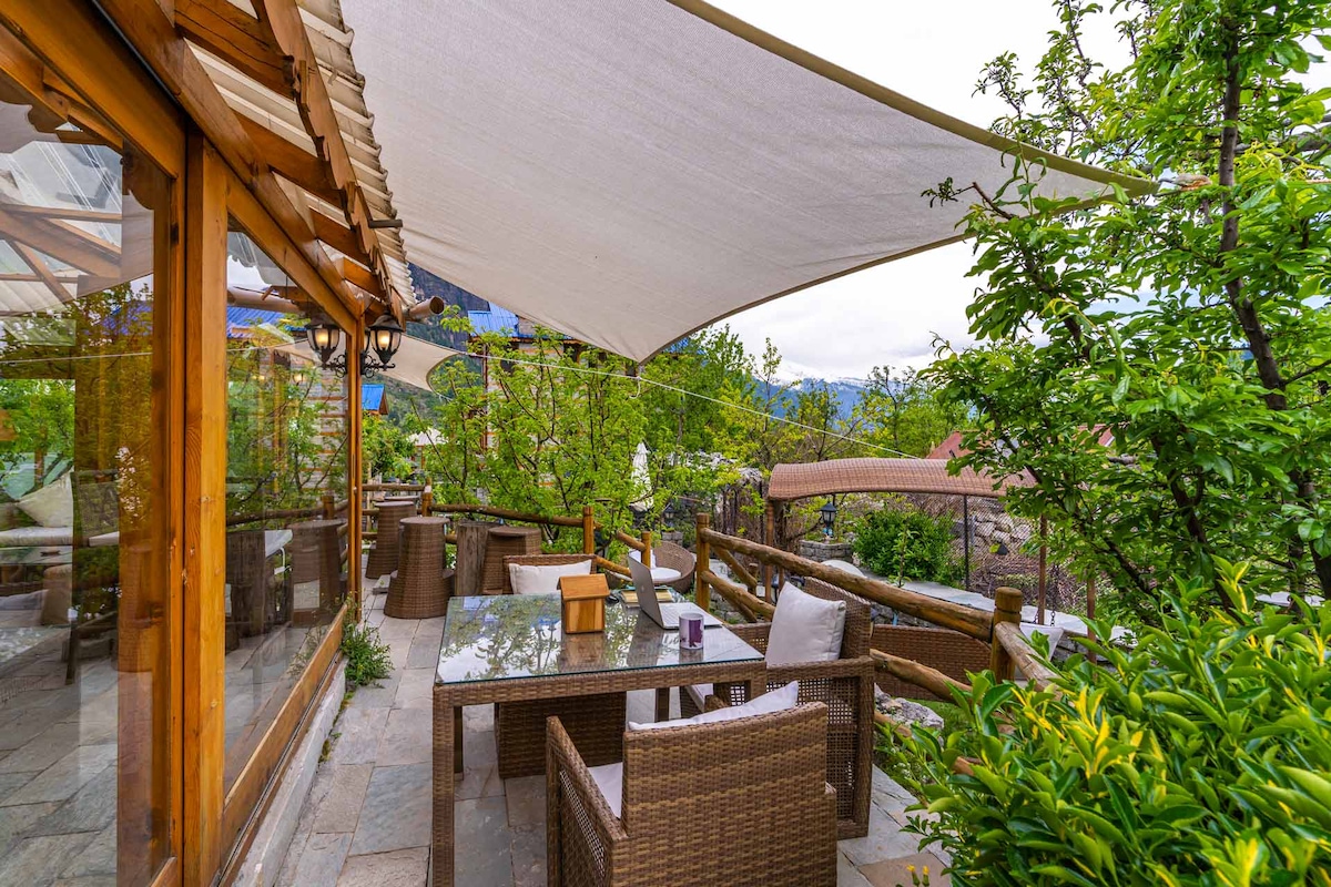 An outdoor dining area features wooden furniture under a protective awning, surrounded by lush greenery and flowering plants. The serene setting invites relaxation, with stone flooring and a natural landscape that complements the rustic charm of the cabin.