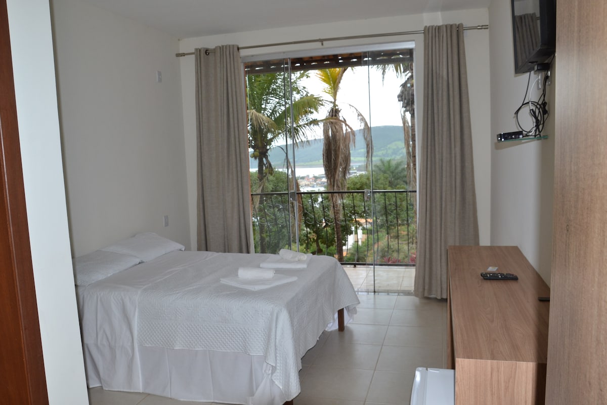 A well-lit room is furnished with two single beds, each dressed in crisp white linens. Floor-to-ceiling windows provide a view of palm trees and a distant lake. A wooden TV stand and air conditioning unit are positioned against the wall, enhancing the room's functionality.