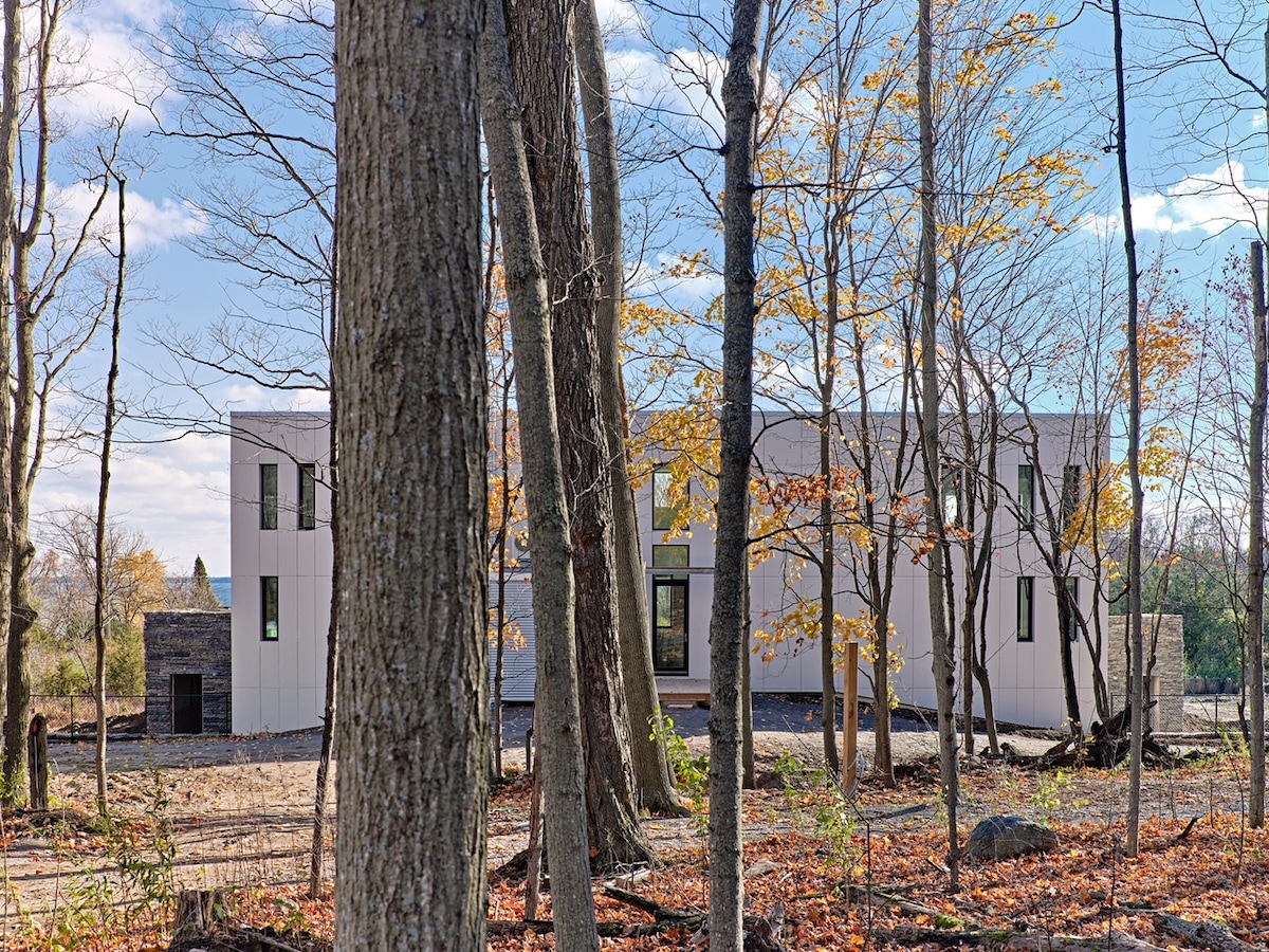 A modern architectural structure is showcased amidst a wooded setting, surrounded by bare trees with hints of autumn foliage. The exterior features a sleek design and multiple large windows, offering a glimpse of the spacious interior. A serene natural environment enhances the scene.