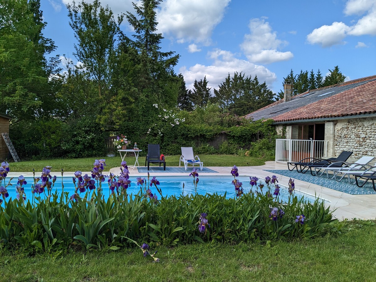 A serene pool area surrounded by blooming irises. Comfortable lounge chairs are positioned around the water's edge. A charming farmhouse with a rustic roof is visible in the background, framed by lush greenery and soft clouds above.