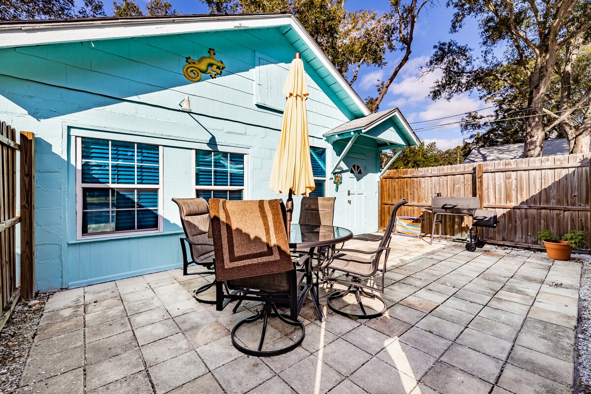 An outdoor patio is set with a circular table surrounded by six chairs. An umbrella provides shade for the seating area, while a grill is positioned against the back wall. A privacy fence and potted plant enhance the space, framed by trees.