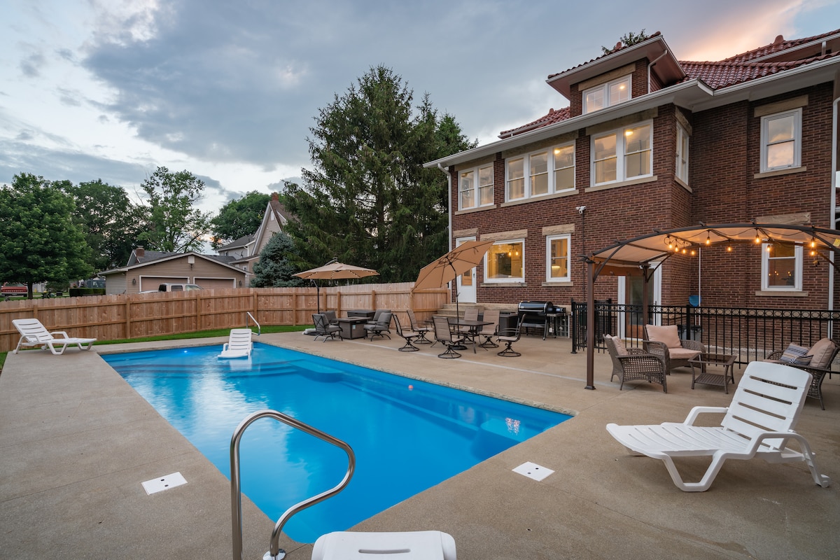 A spacious outdoor area features a pool surrounded by lounge chairs and shaded seating under umbrellas. The two-story brick house stands in the background, with trees and neighboring homes visible beyond the pool area. Warm lighting from string lights enhances the ambiance.