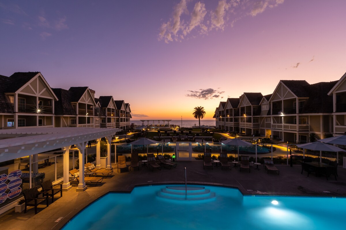 The image captures a resort at twilight, showcasing a tranquil pool with a glowing blue hue. The backdrop consists of elegant buildings with peaks, harmonizing beautifully with the sunset sky, which transitions from purple to orange. Palm trees are visible near the ocean horizon.