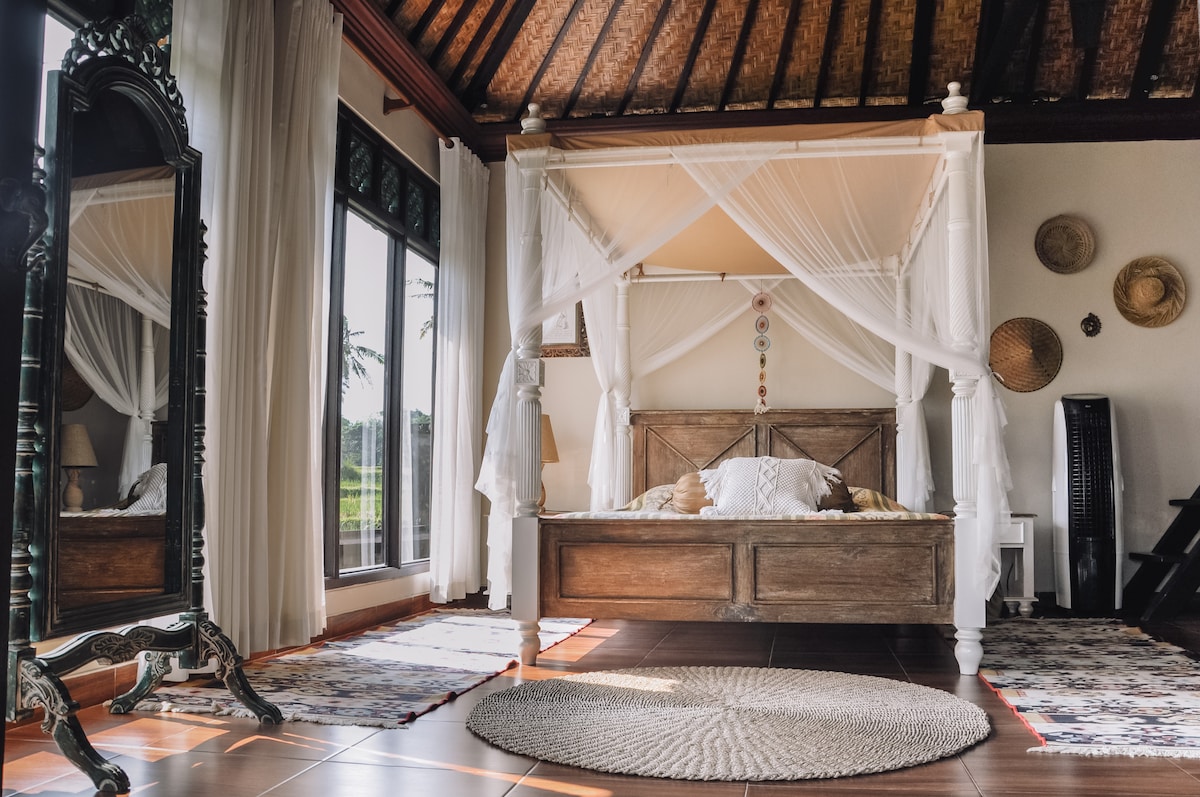 A serene bedroom features a wooden four-poster bed draped with sheer white fabric. Large glass doors allow natural light to fill the space, while a round rug lies on the tiled floor. Decorative elements include woven hats on the wall and a full-length mirror.