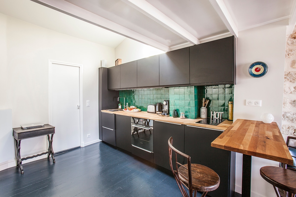 The kitchen features sleek black cabinetry with a wooden countertop, highlighted by a vibrant green tiled backsplash. A table with two bar stools is present, alongside modern appliances including a kettle and coffee maker. A door leads to another space, maintaining a clean and organized environment.