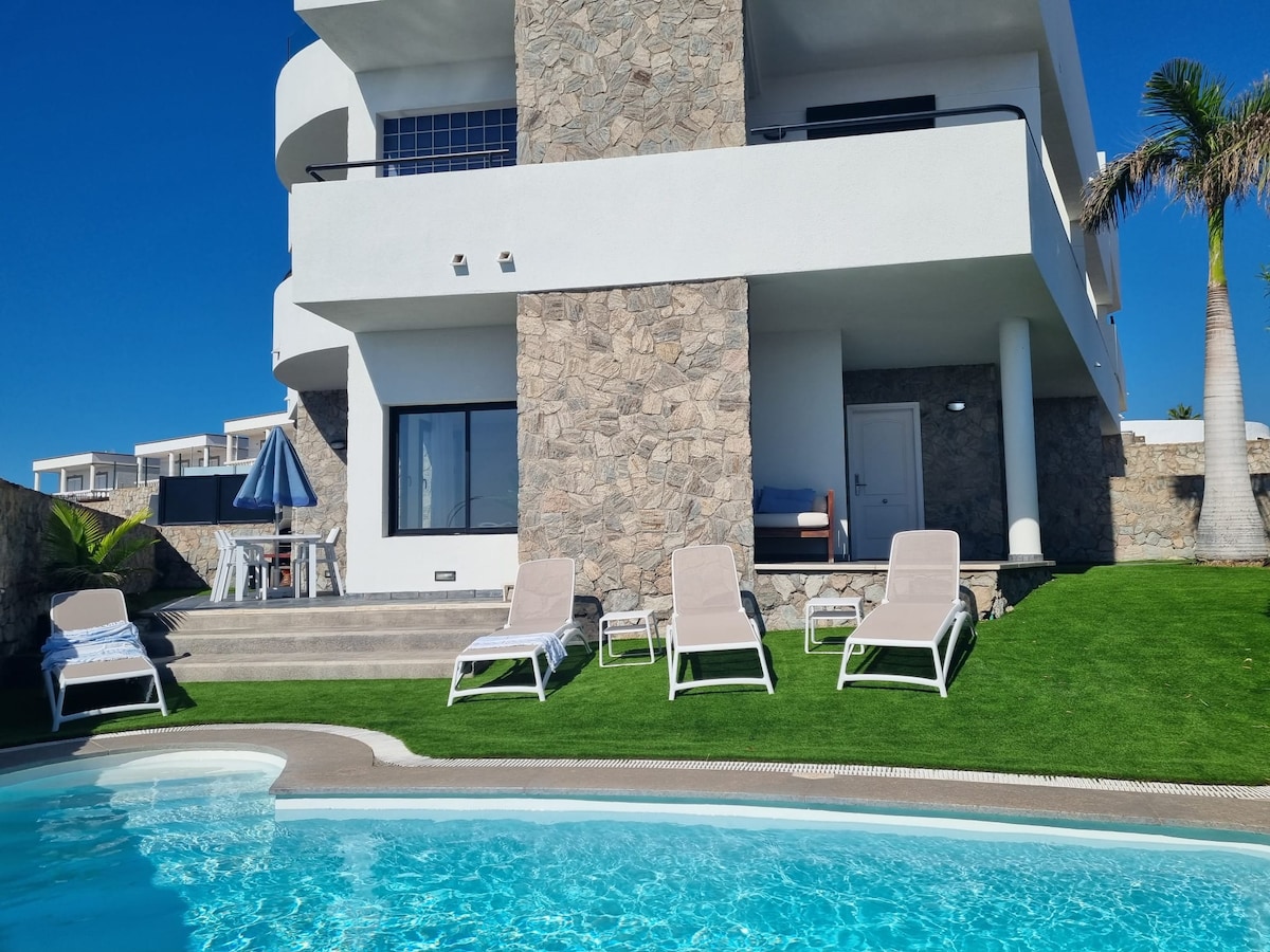 The villa presents a modern exterior with smooth white walls and stone accents. A private pool glistens beside a manicured lawn, while several loungers offer a place to relax. A shaded seating area is visible, enhancing the inviting outdoor space.