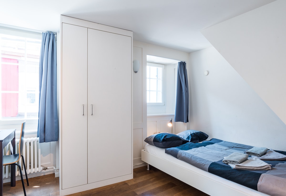 A well-lit sleeping area features a spacious bed with neatly arranged linens. Natural light enters through a window, complemented by light curtains. A white wardrobe occupies one corner, while a small desk chair is positioned nearby, contributing to the functional layout.