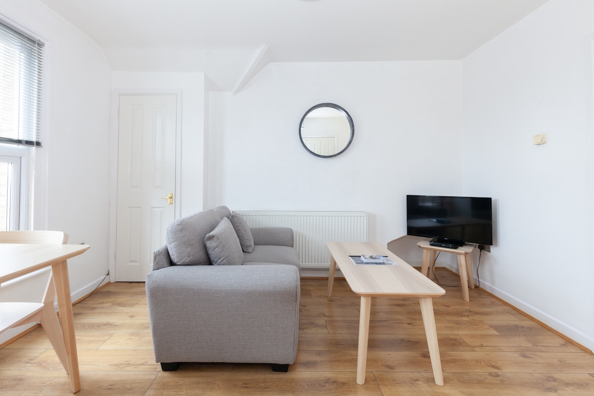 A light-filled living area is furnished with a gray sofa and a wooden coffee table. A round mirror is mounted on the wall opposite, and a small television is situated on a corner stand. Neutral tones and wooden flooring contribute to a clean, modern aesthetic.