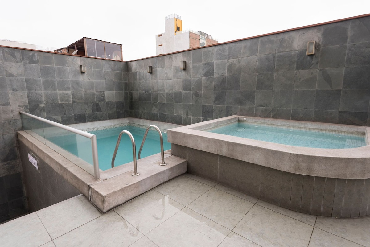 A pool area features two distinct water sections, one rectangular and the other round, each with smooth stone finishes. The space is enclosed by textured stone walls, creating a serene environment, and the floor is covered in large, light-colored tiles, contributing to a modern aesthetic.