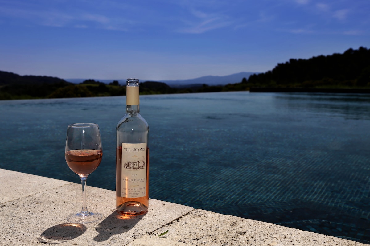 A glass of rosé wine sits beside an open bottle on the edge of a tranquil infinity pool. The water reflects the clear blue sky and surrounding hills, creating a serene atmosphere. Lush greenery frames the scene in the background.