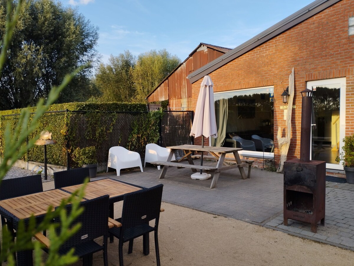 A furnished terrace area features a wooden picnic table and chairs, alongside a firepit. An umbrella provides shade. The backdrop includes a brick building with large glass doors, complemented by greenery and a gravel pathway.