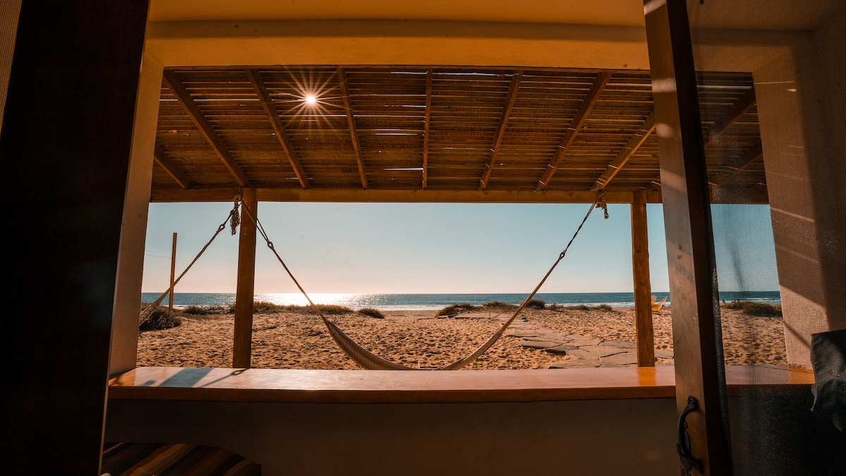 A serene view of a beachfront terrace features a hammock swaying gently, positioned under a bamboo roof. Sunlight reflects off the calm waves of the ocean, creating a peaceful ambiance that invites relaxation and enjoyment of the natural surroundings.