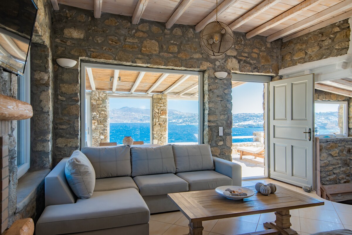 A living area presents a spacious light-colored sofa set against a stone wall, providing a cozy spot with a view of the Aegean Sea. Large glass doors open to a sunlit terrace, enhancing the connection to the outdoors. A wooden coffee table is neatly arranged with decorative items.
