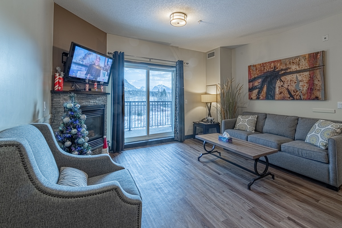 The living area features a cozy gray sofa and an armchair, complemented by a modern wooden coffee table. A flat-screen TV is positioned opposite a large glass door leading to a private balcony. The space is illuminated by soft lighting and offers a view of the mountains.