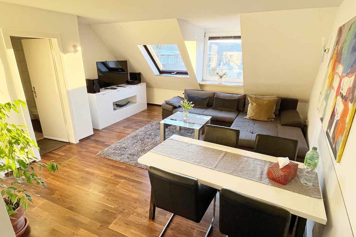 The living area features a spacious grey couch accompanied by modern furniture. A glass coffee table sits on a plush rug, while a dining table with black chairs is positioned nearby. Natural light enters through angled windows, enhancing the bright and welcoming environment.