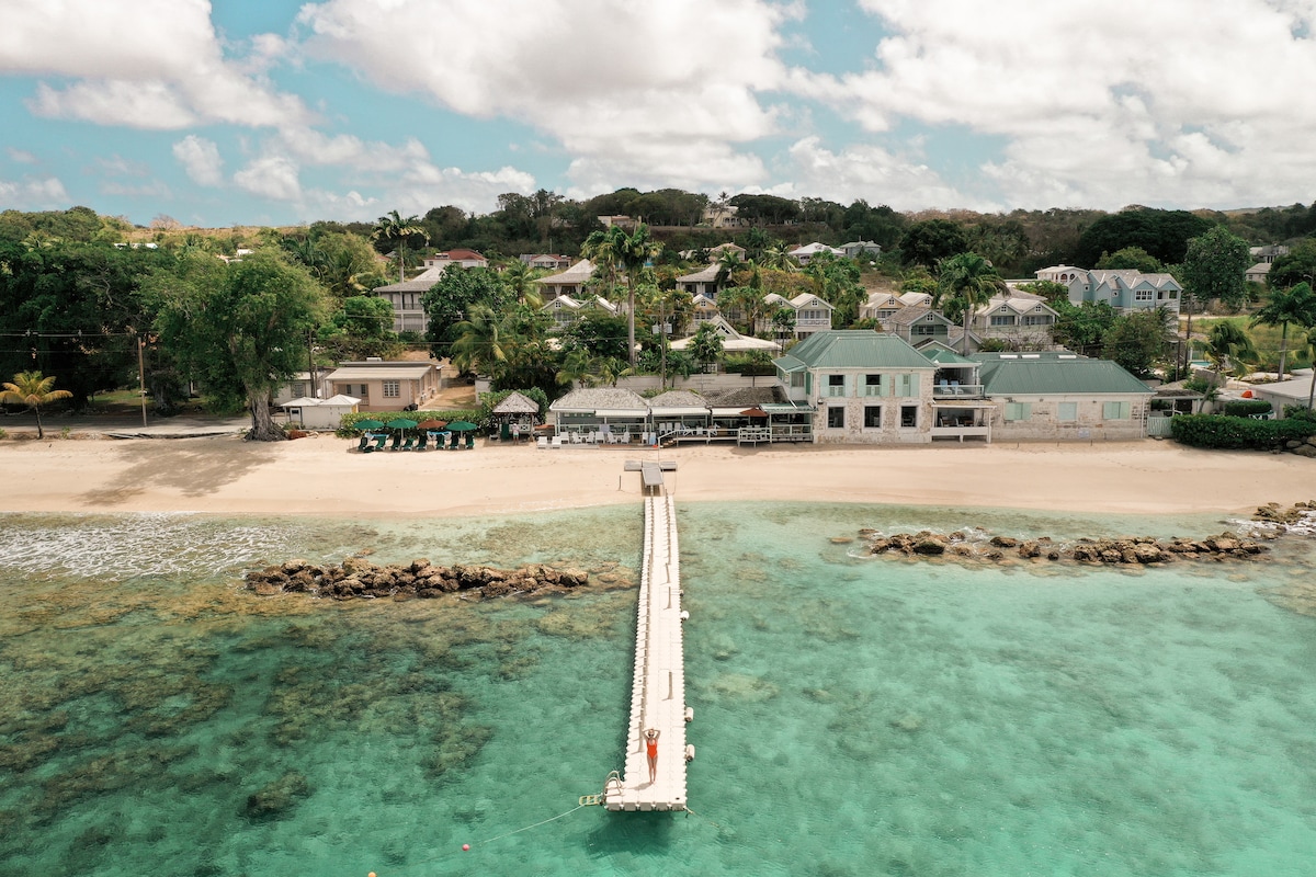 An aerial view captures a wooden pier extending into turquoise waters, leading to a sandy beach. The shoreline is lined with lush greenery and beachside structures, offering a relaxed coastal atmosphere. The scene is framed by a vibrant tropical landscape under a partly cloudy sky.