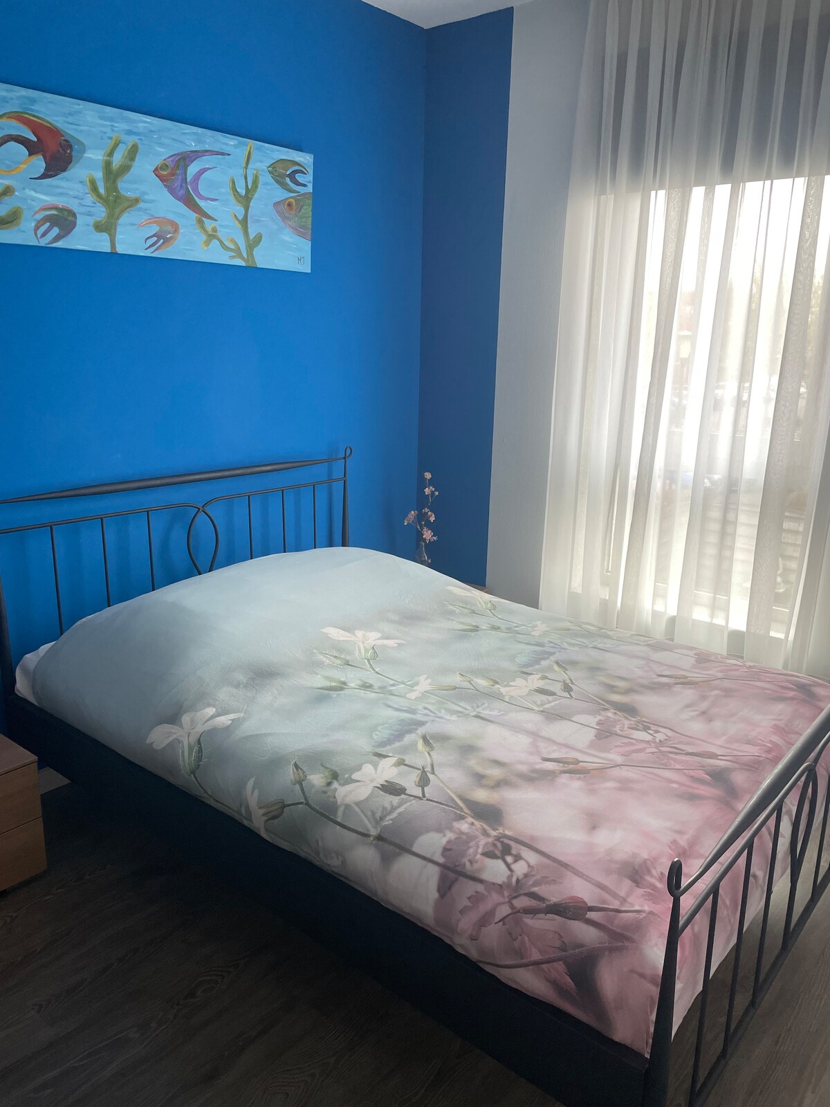 A sleeping area features a dark metal bedframe with a floral blanket covering the spacious bed. The vibrant blue walls are complemented by a piece of wall art and ample natural light filtering through sheer white curtains. A bedside table is positioned next to the bed.