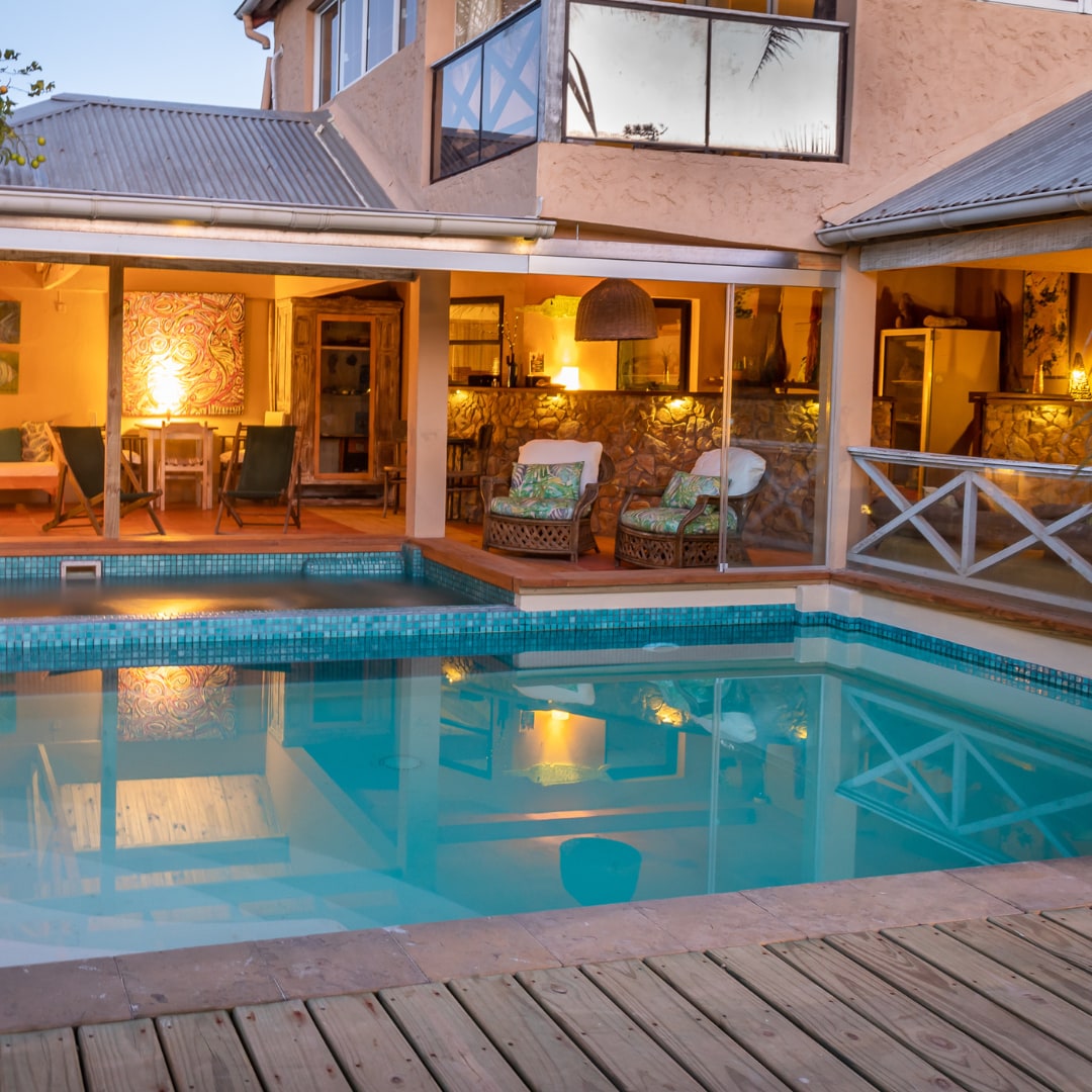 An outdoor area features a rectangular pool surrounded by a wooden deck. Comfortable seating options, including two lounge chairs, are positioned near the pool. The warm glow of interior lights from the adjacent living space creates a welcoming ambiance, reflected on the water's surface.