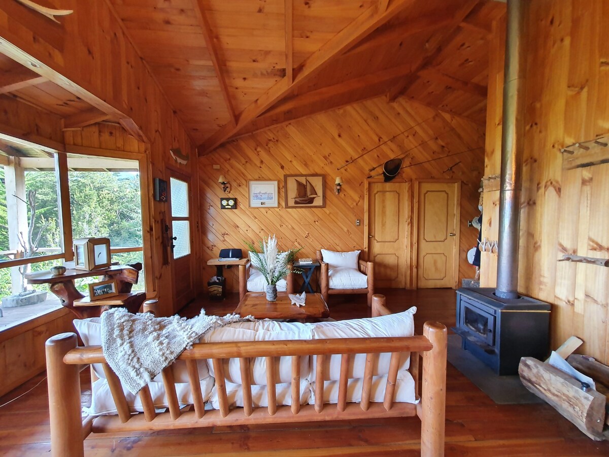 A wooden interior is featured, showcasing a cozy sitting area adorned with light-colored furniture. Large windows allow natural light to fill the space, with views of greenery visible outside. A rustic wood-burning stove adds warmth, complemented by wooden beams and wall decor.