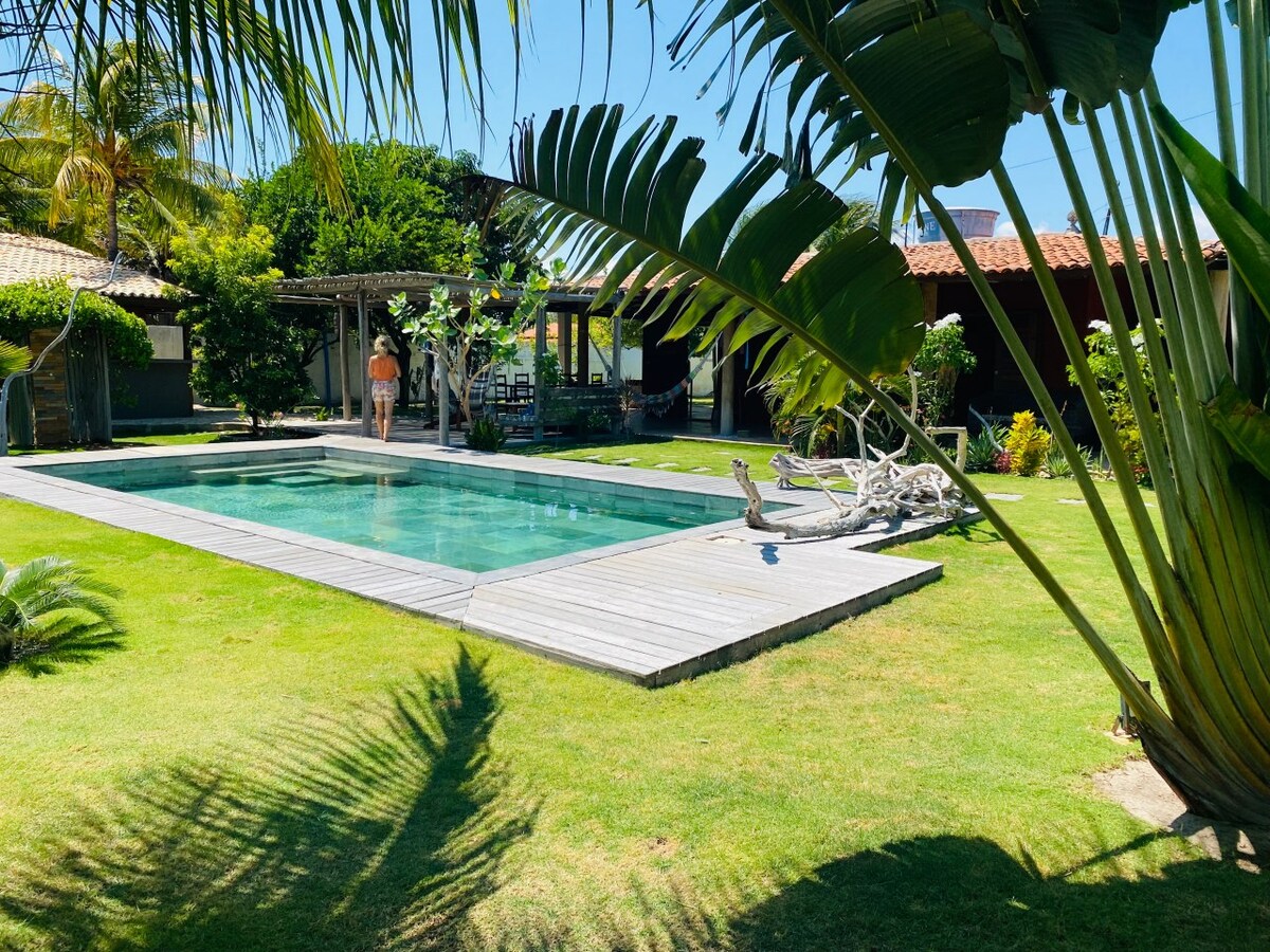 A natural stone pool is framed by a wooden deck and surrounded by vibrant greenery. The open outdoor area features lounge chairs and a partially shaded patio with a clear view of the house in the background.