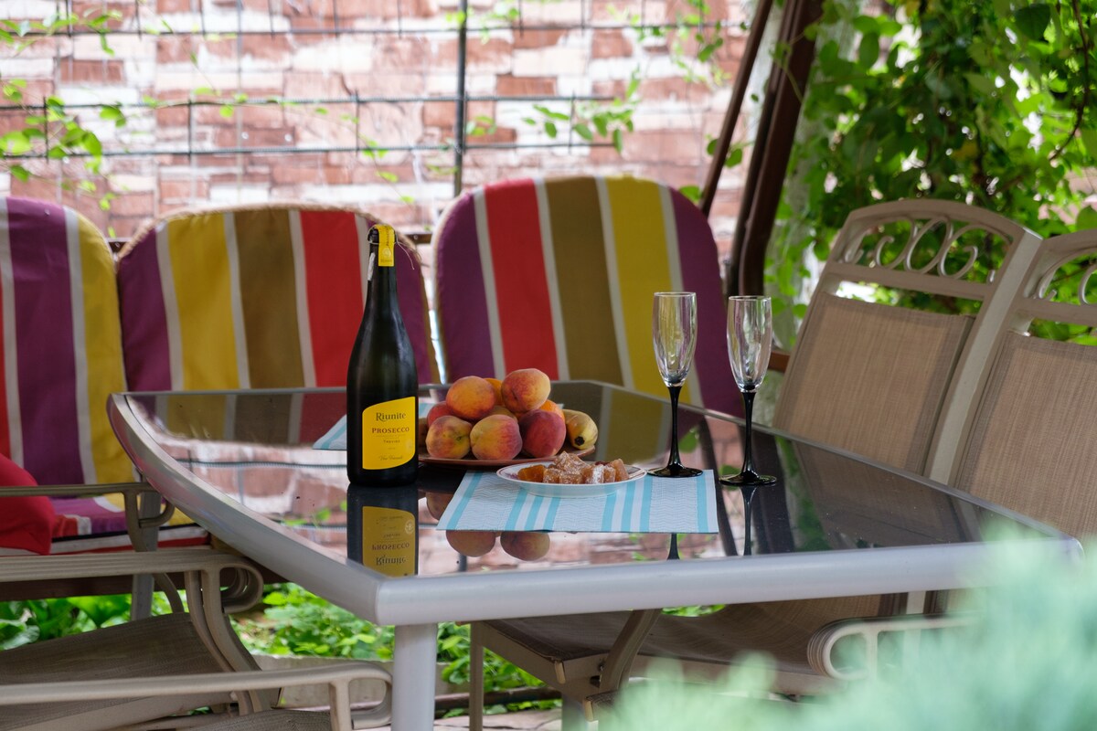 An outdoor dining area is set with a glass table and several chairs featuring colorful striped cushions. A bottle of sparkling wine sits alongside glasses, with fresh peaches displayed on a plate. Lush greenery provides a natural backdrop, enhancing the space.