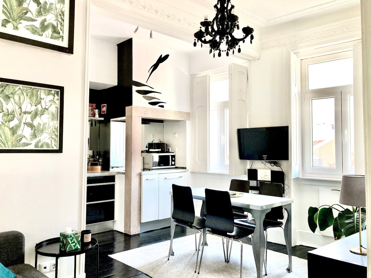 A modern living space features a dining table with black chairs, complemented by a bright kitchenette visible in the background. Large windows allow natural light to fill the room, and a chandelier adds an elegant touch. Decorative wall art incorporates leafy designs.