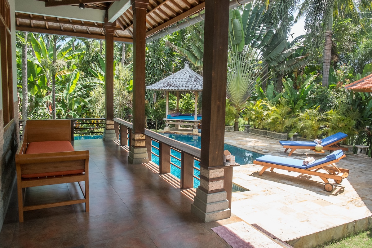 An inviting terrace area showcases comfortable seating alongside a tranquil pool. Sun loungers are positioned near the water, surrounded by lush greenery and palm trees, creating a serene outdoor space that accentuates relaxation and leisure.