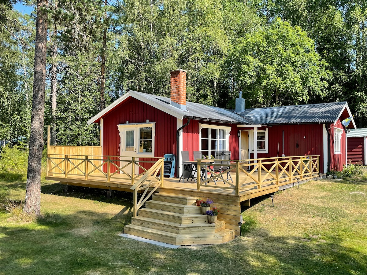 A charming red cottage is shown with a wooden deck and surrounding greenery. The deck features outdoor seating and a small table with potted flowers. A pathway of steps leads up to the entrance, enhancing accessibility.