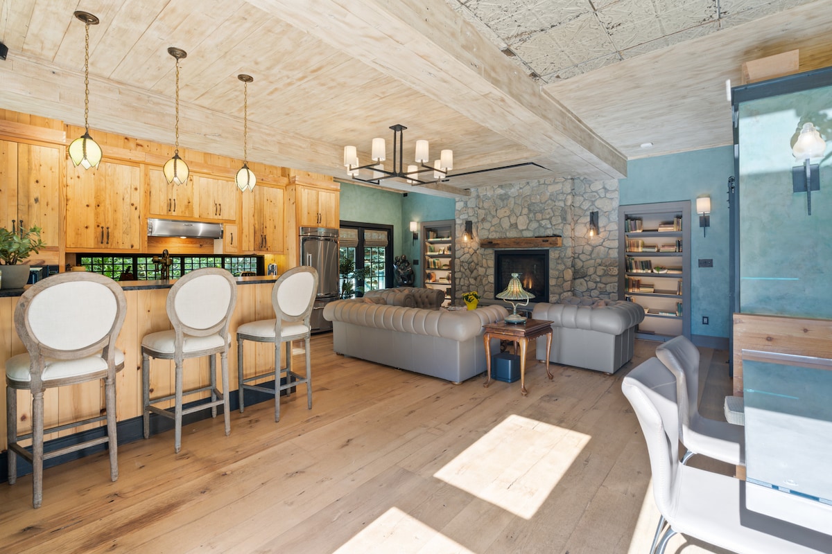 An open living space features a modern kitchen with a large island and sleek bar stool seating. A cozy stone fireplace is positioned in the lounge area, surrounded by comfortable couches. Warm wooden accents and natural light enhance the room's inviting atmosphere.