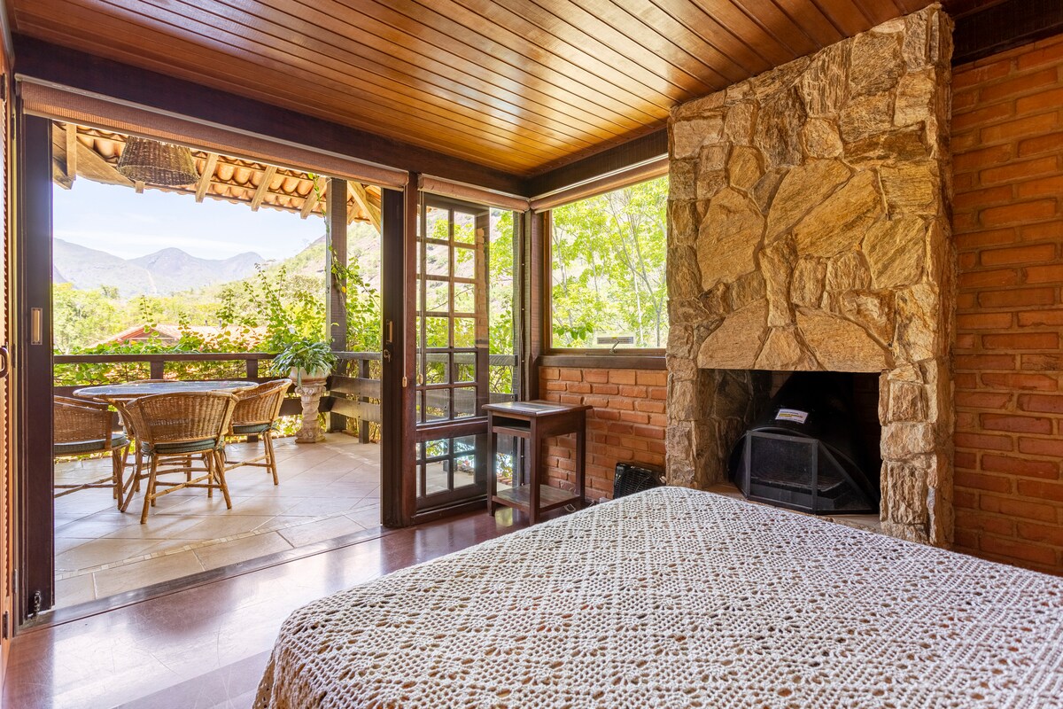 A cozy bedroom features a spacious bed adorned with a patterned blanket. Large glass doors open to a balcony with a wooden table and chairs, offering views of the surrounding nature. A stone fireplace adds a rustic charm to the room.