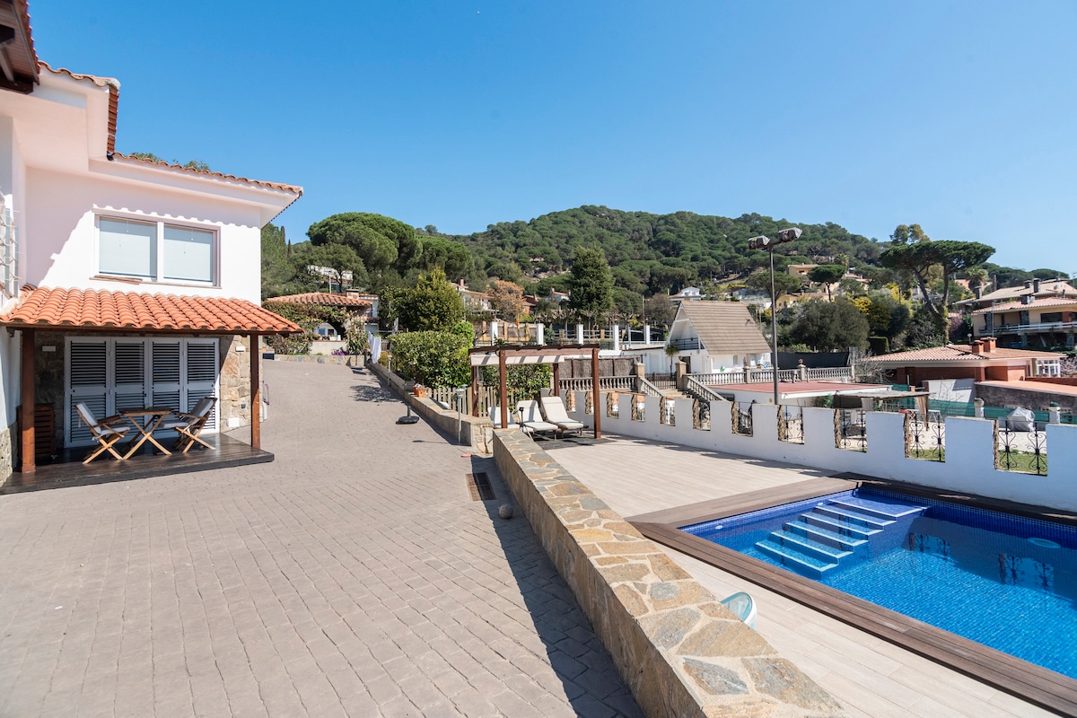 An outdoor space featuring a pool bordered by a wooden deck and loungers. The view showcases surrounding green hills and residential areas, with an inviting path leading to the house, which has a terracotta roof and outdoor seating.