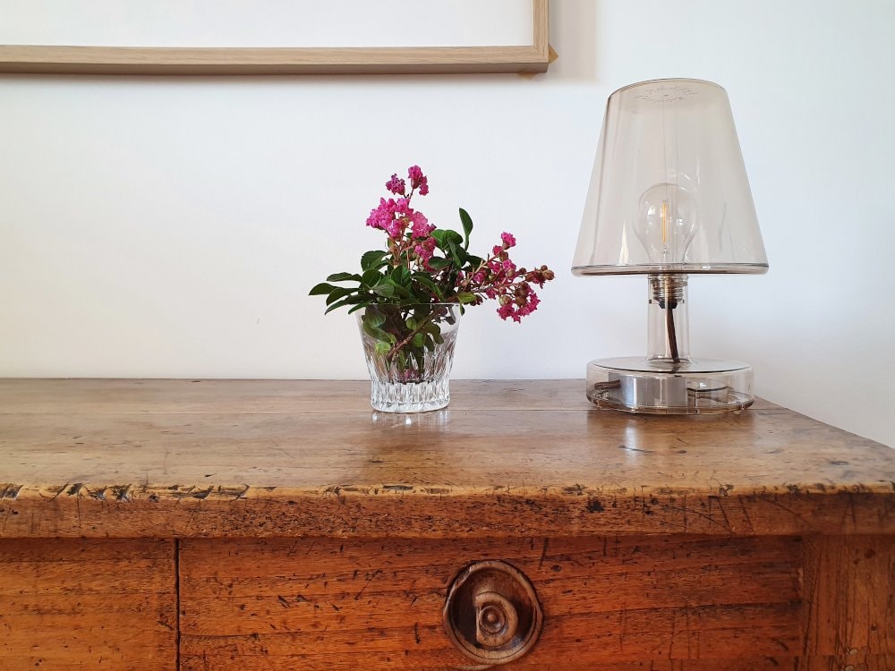 A wooden sideboard features a small vase containing pink flowers alongside a stylish glass lamp. The textured surface of the sideboard highlights its rustic charm, complemented by the clean, neutral wall in the background.
