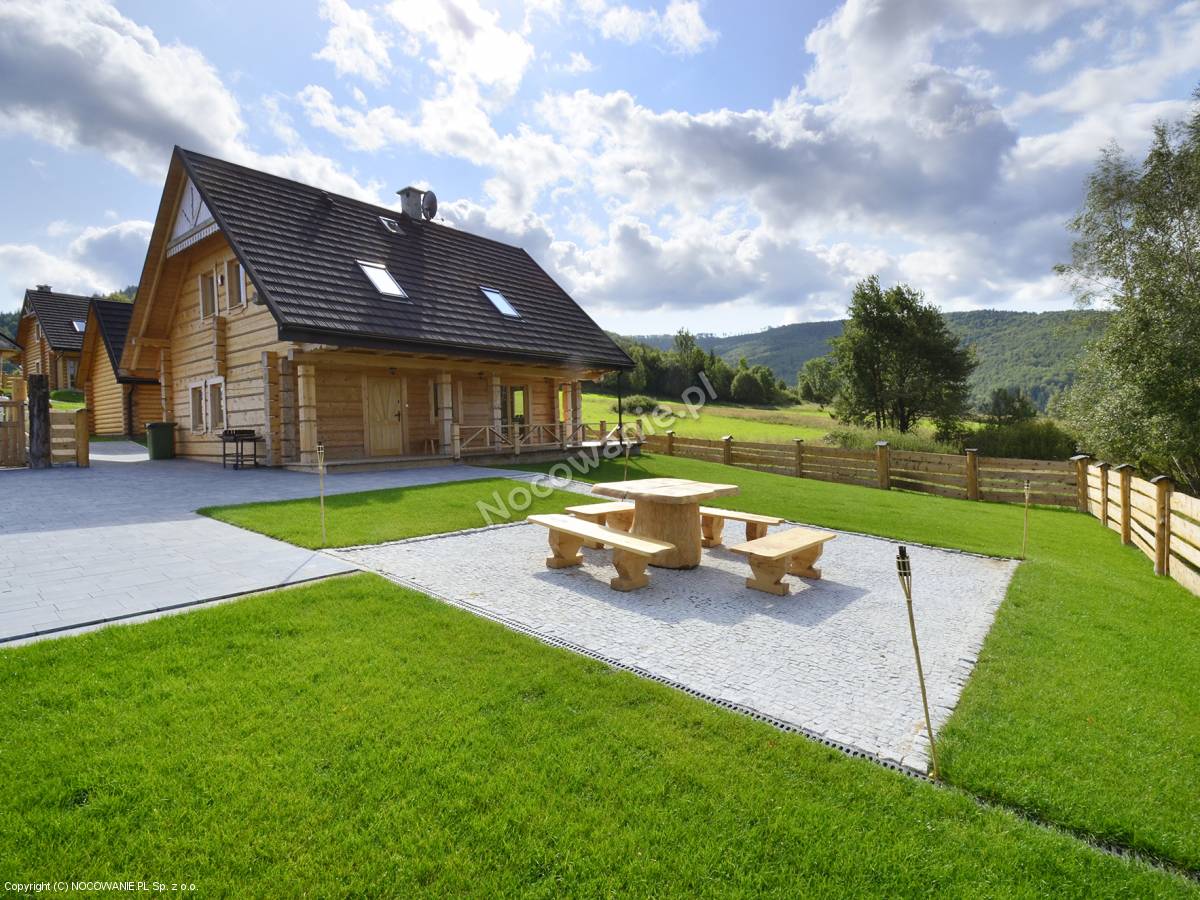 A spacious outdoor area features a circular wooden table surrounded by benches, set on a stone patio. Lush green grass surrounds the space, complemented by a wooden fence and distant hills under a partly cloudy sky.