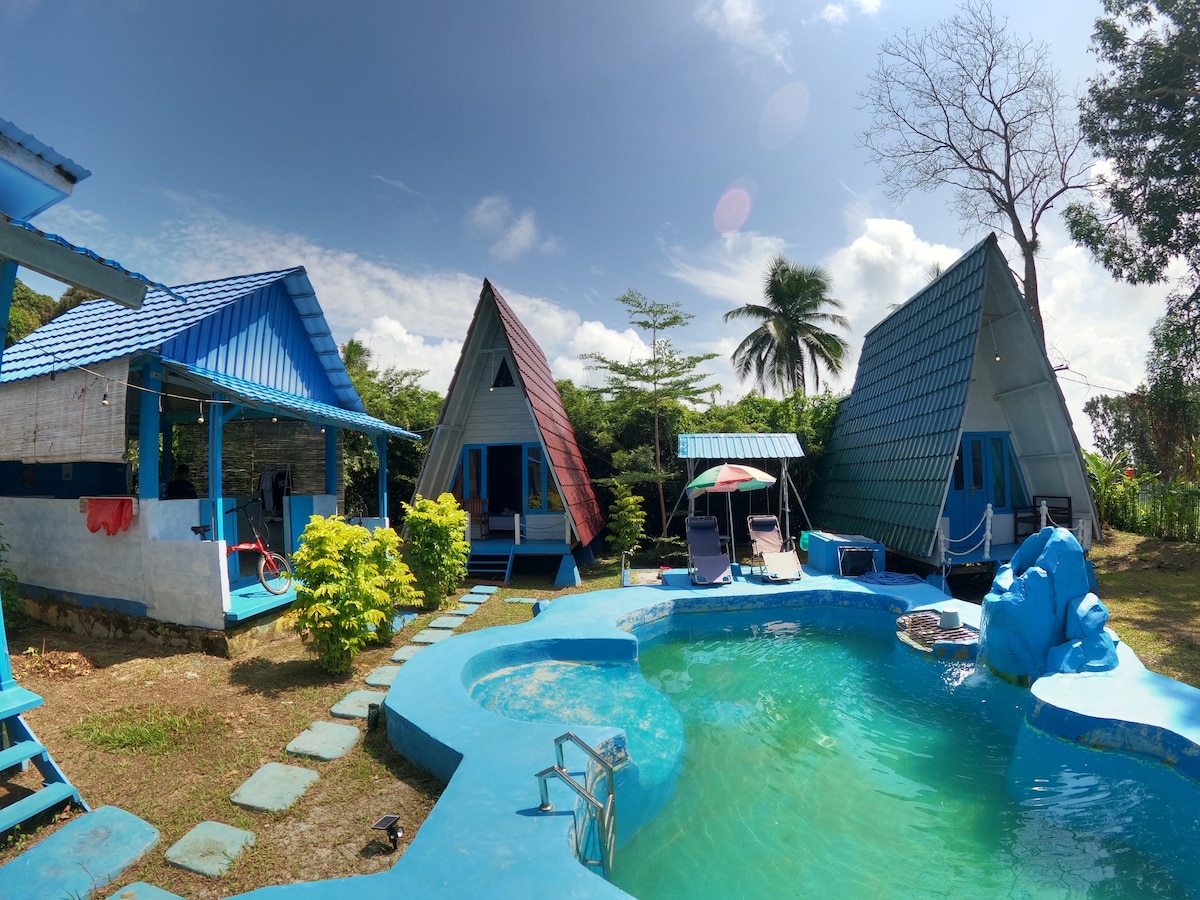Three A-frame cottages are arranged around a turquoise pool. The pool area is complemented by vibrant greenery and sun loungers. Each cottage features a distinct roof color, and palm trees are visible in the background against a clear blue sky.