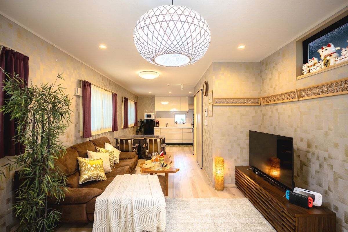 A spacious living area is highlighted by a modern chandelier and a large flat-screen TV. A comfortable brown sofa sits in front of a wooden coffee table, adorned with decorative items. Light streams in through windows framed by deep burgundy curtains, illuminating the space.