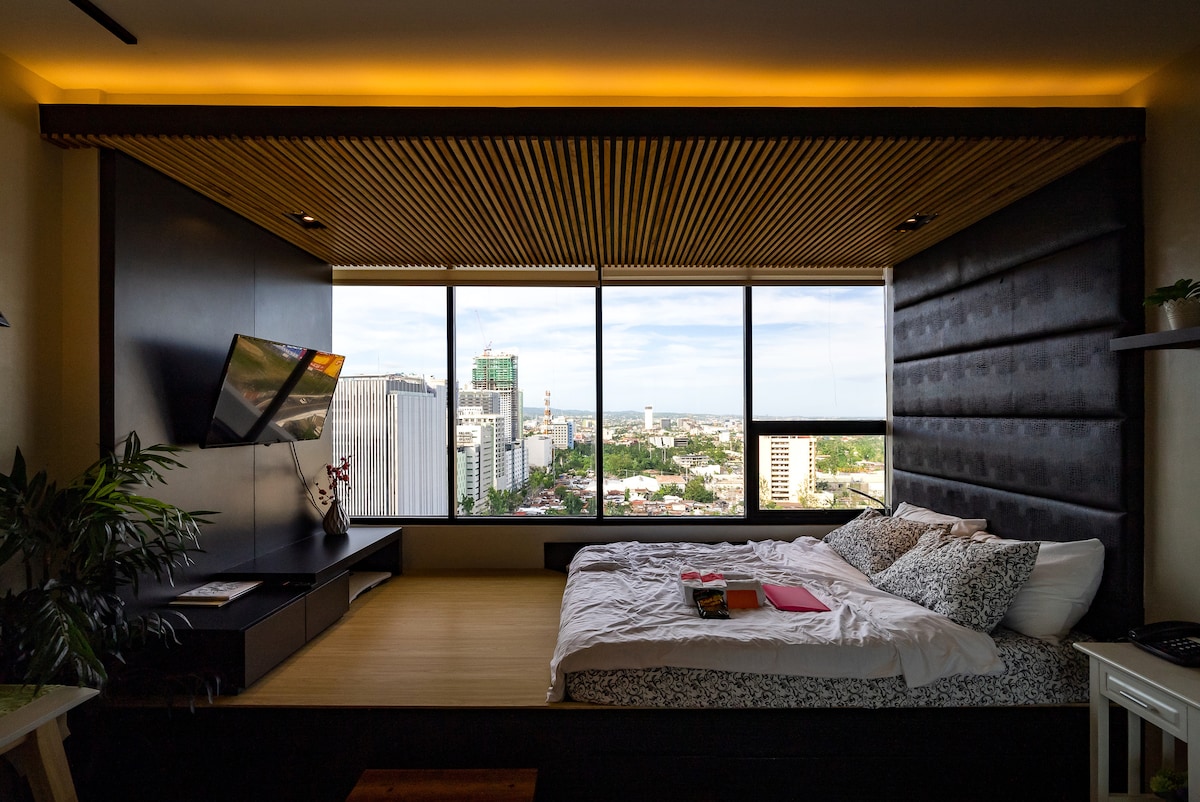 A contemporary studio bedroom features a spacious bed with soft bedding, positioned by large windows that offer panoramic city views. A flat-screen TV is mounted on the wall opposite the bed, with wooden ceiling accents adding warmth to the modern design.