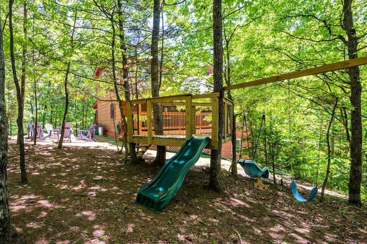 A play area is nestled among trees, featuring a slide, swings, and a climbing structure. The cabin is visible in the background, surrounded by greenery. The inviting outdoor space offers a natural setting for children to play, enhancing the rental's family-friendly appeal.