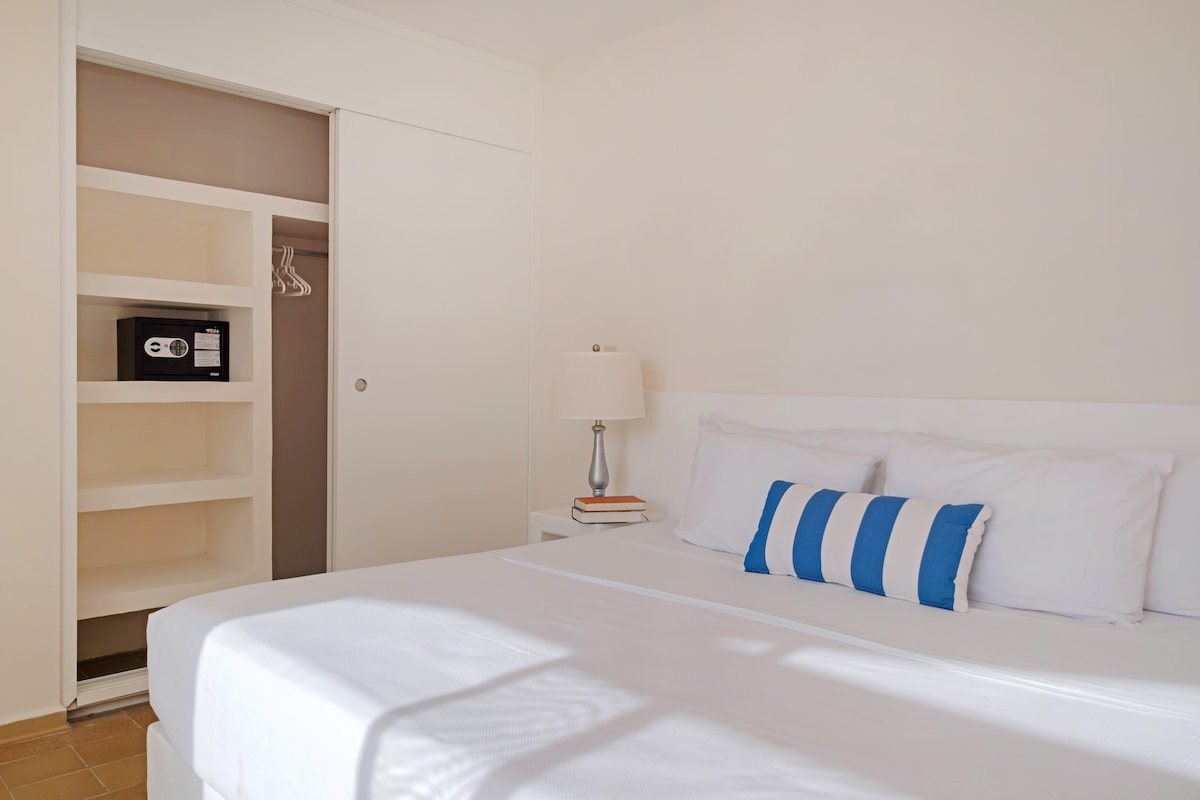 A well-lit sleeping area includes a spacious bed with white linens and decorative striped pillows. A nightstand with a lamp sits beside the bed. An accessible closet features shelving and a safe, adding to the suite's convenience.