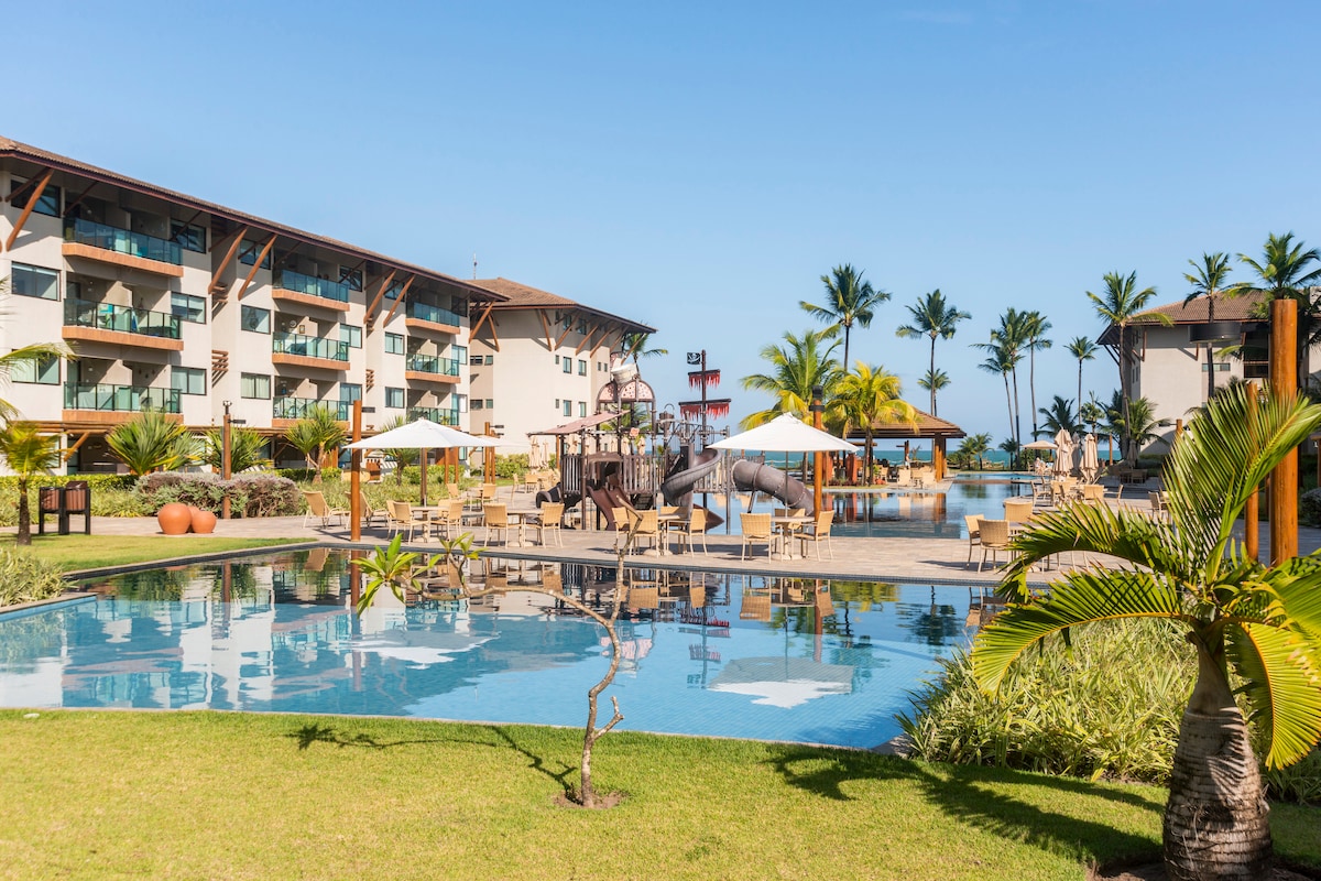 The landscaped area features a large swimming pool surrounded by tropical palm trees. Sun loungers with umbrellas are positioned around the pool, while a water slide and playground are visible in the background, offering recreational options for families.