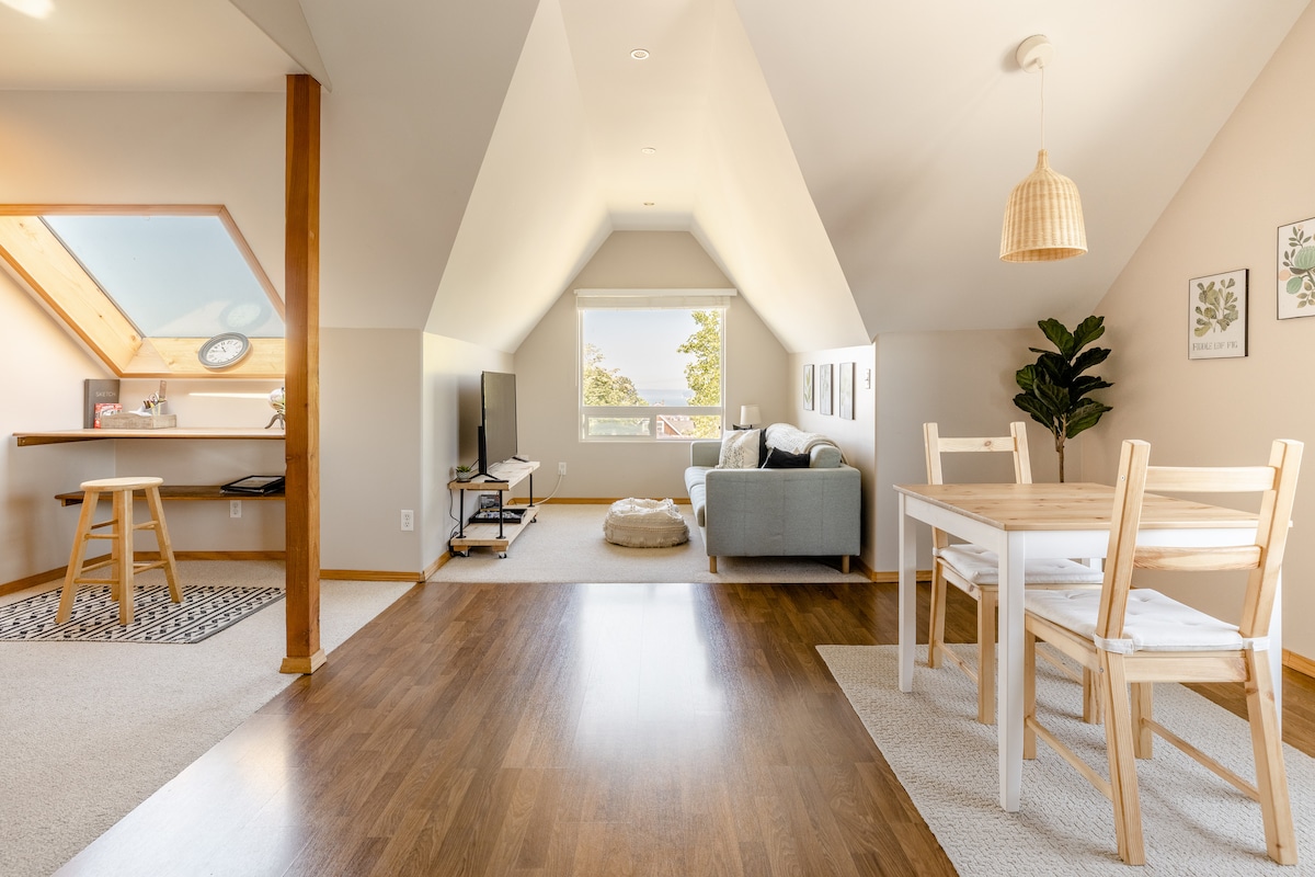 The airy living area features sloped ceilings and ample natural light, creating a bright atmosphere. A comfortable sofa invites relaxation, while a dining table and chairs sit nearby. A small kitchenette is visible in the background, complemented by decorative plants and artwork on the walls.