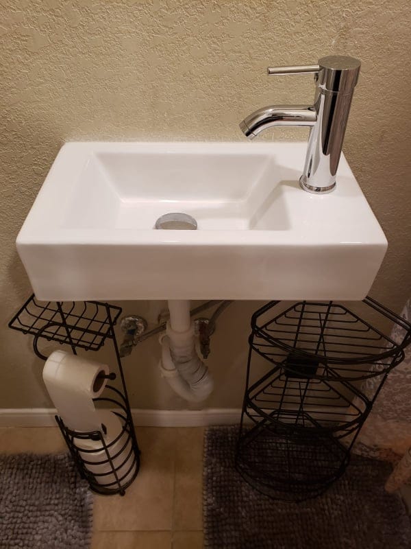 A compact sink is positioned above a sleek, modern basin with a chrome faucet. The structure is flanked by two black wire racks, providing convenient storage for toiletries and toilet paper. The surrounding wall features a light, textured finish.