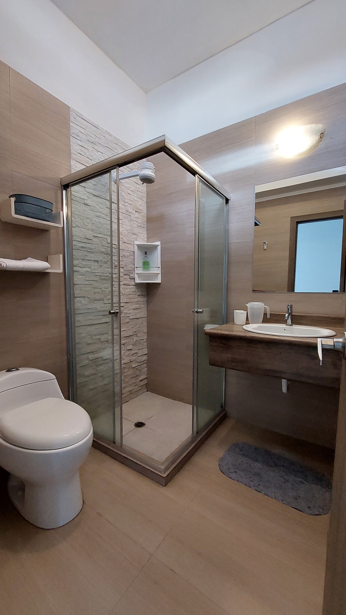 A modern bathroom is equipped with a glass-enclosed shower featuring stone accents. A wooden vanity with a wash basin and toiletries sits alongside a toilet. Soft lighting provides a calming ambiance, and a small rug is placed on the floor for comfort.