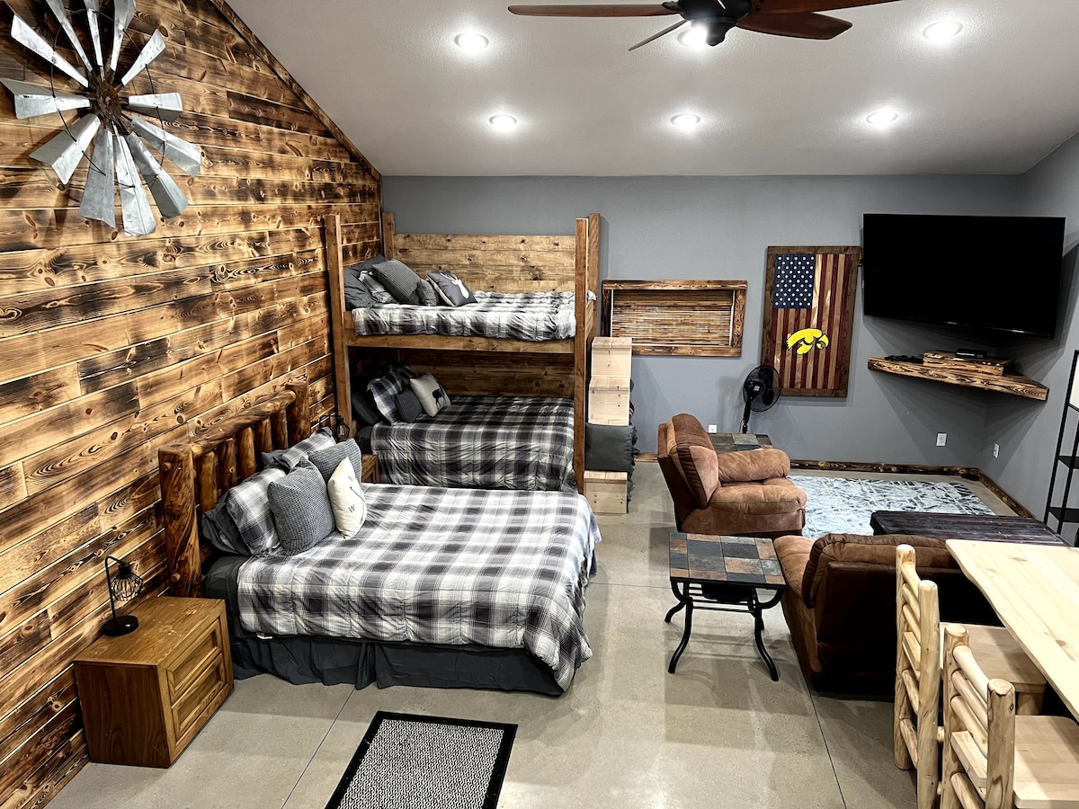 A spacious living area features rustic wood accents and cozy furnishings. A bunk bed and a spacious bed are adorned with plaid bedding. A comfortable recliner and a small dining table complete the space, with a flat-screen TV mounted on the wall.