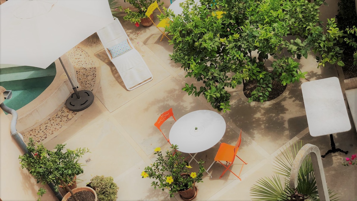 A sunny courtyard is seen with lush greenery and potted plants. A white table is surrounded by bright orange chairs. A white umbrella provides shade over a lounge chair, complementing the peaceful outdoor space.